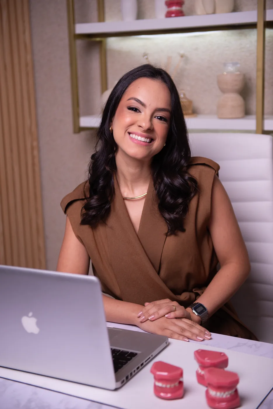 Dra. Rafaela Silva sorrindo em sua mesa, com modelos de dentes usados para explicar a higiene bucal às crianças